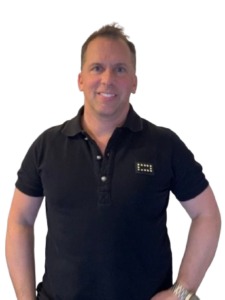 Porträt eines selbstbewussten Mannes im schwarzen Poloshirt vor dunklem Hintergrund, freundliches Lächeln, stehende Pose mit Händen an der Hüfte, professionelles Business Profilfoto für Online Marketing, Coaching und Unternehmer Personal Branding.